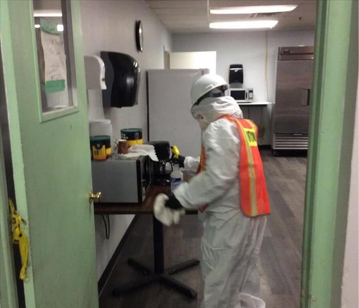 a technician wearing a white Tyvek, gloves, mask, and hardhat cleaning microwave